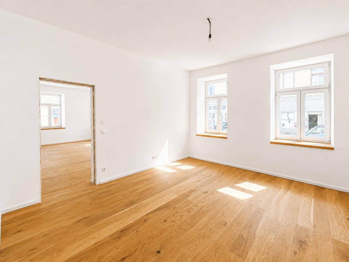 Lichtdurchflutetes Zimmer mit Holzboden und offenem Durchgang zu einem weiteren Raum.