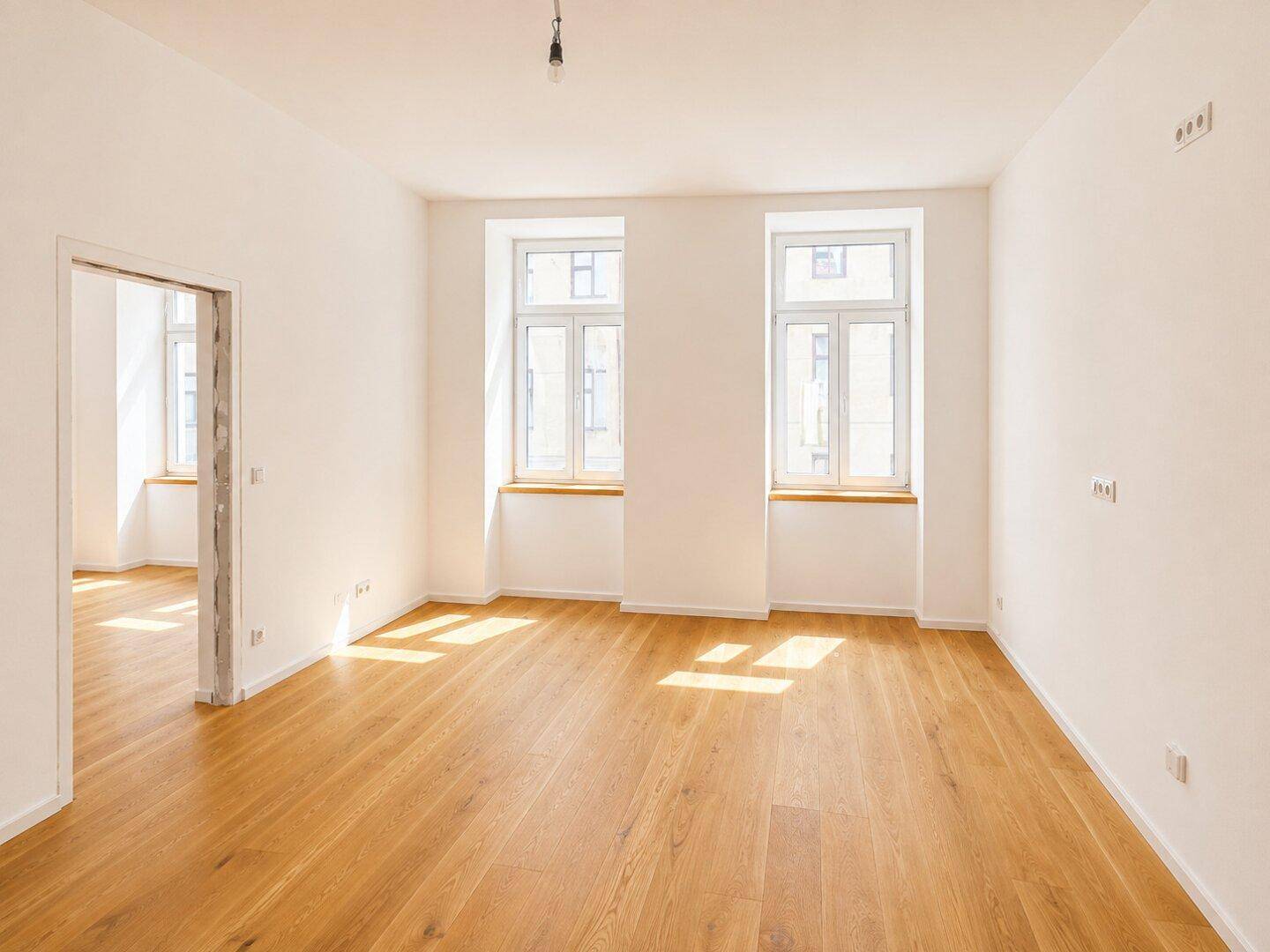 Sonniger, leerer Raum mit schönem Parkettboden und hohen Fenstern, die den Raum erhellen.