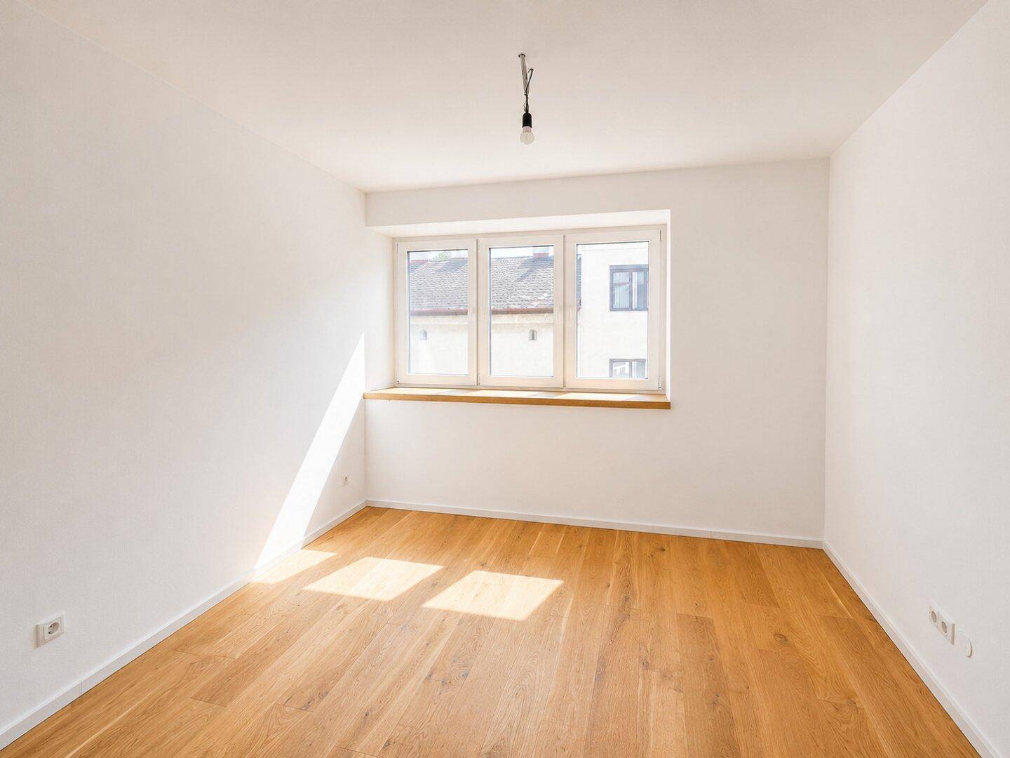 Geräumiges Zimmer mit Holzboden und einem großen Fenster, das den Raum hell erscheinen lässt.