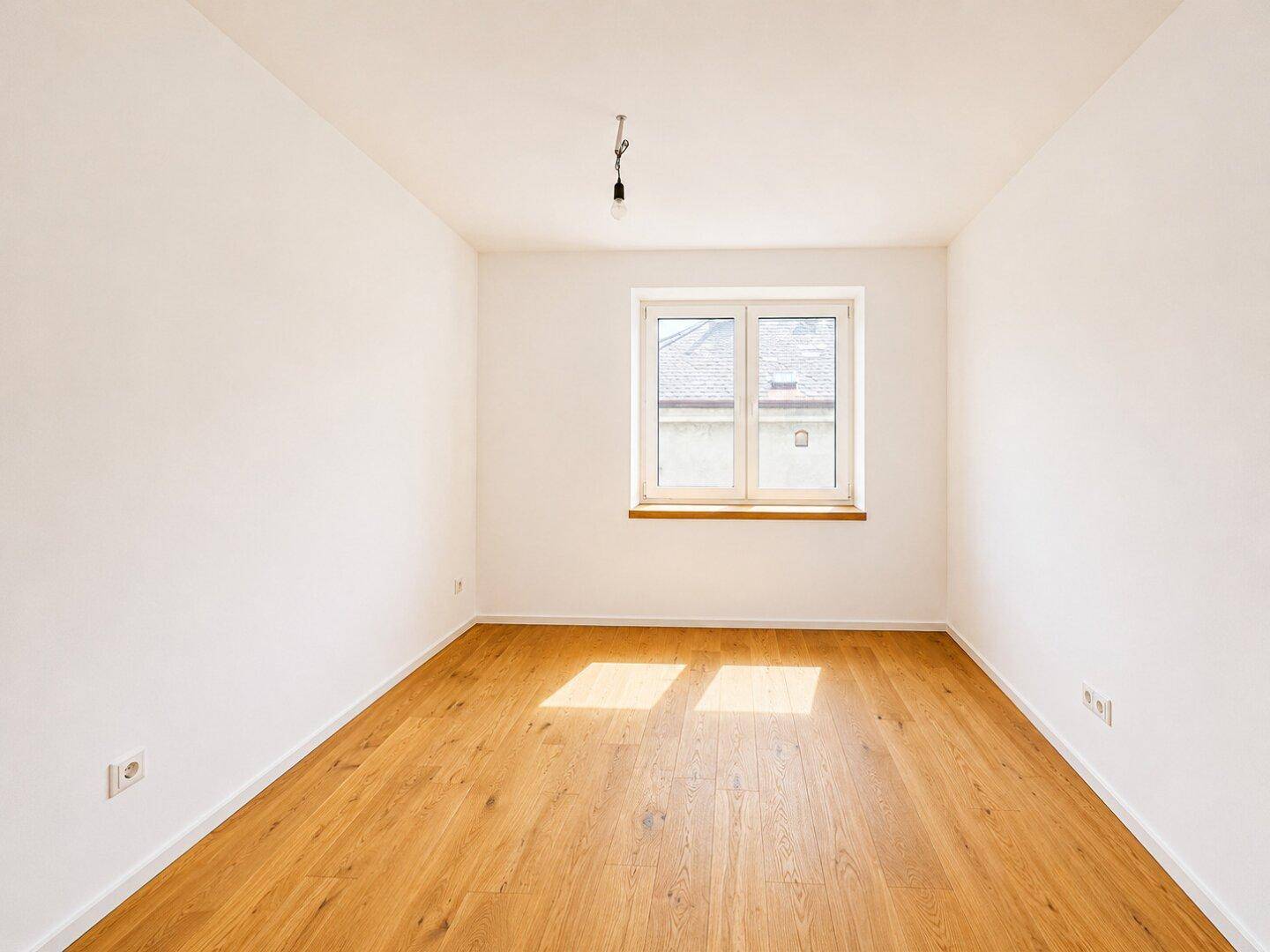 Einfaches, helles Zimmer mit Holzboden und einem Fenster, das eine angenehme Atmosphäre schafft.