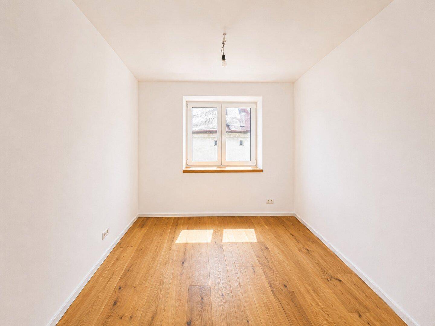 Ruhiges, helles Zimmer mit Holzboden und einem Fenster, das viel Tageslicht hereinlässt.