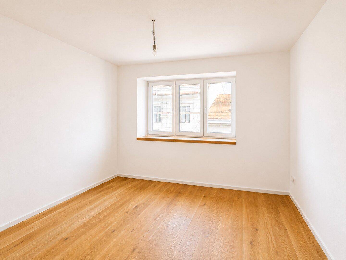 Helles Zimmer mit Holzboden und einem großen Fenster, ideal als Schlaf- oder Arbeitszimmer.