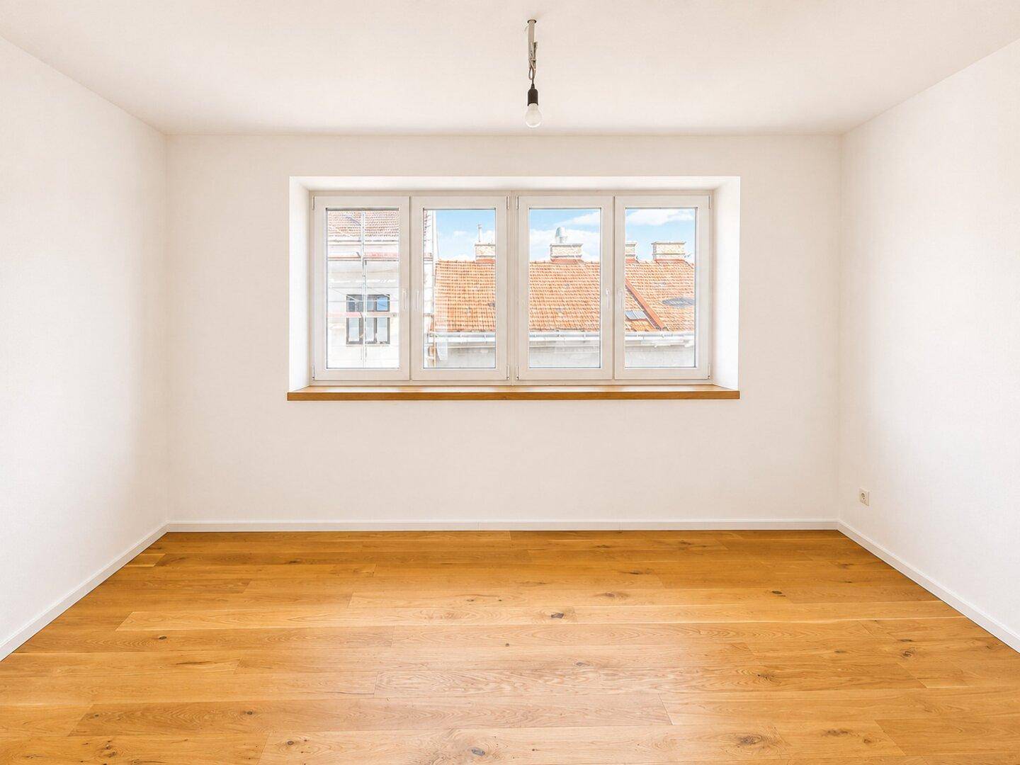 Leerer Raum mit hellem Holzboden und einem Fenster, das einen Blick auf die Dächer bietet.