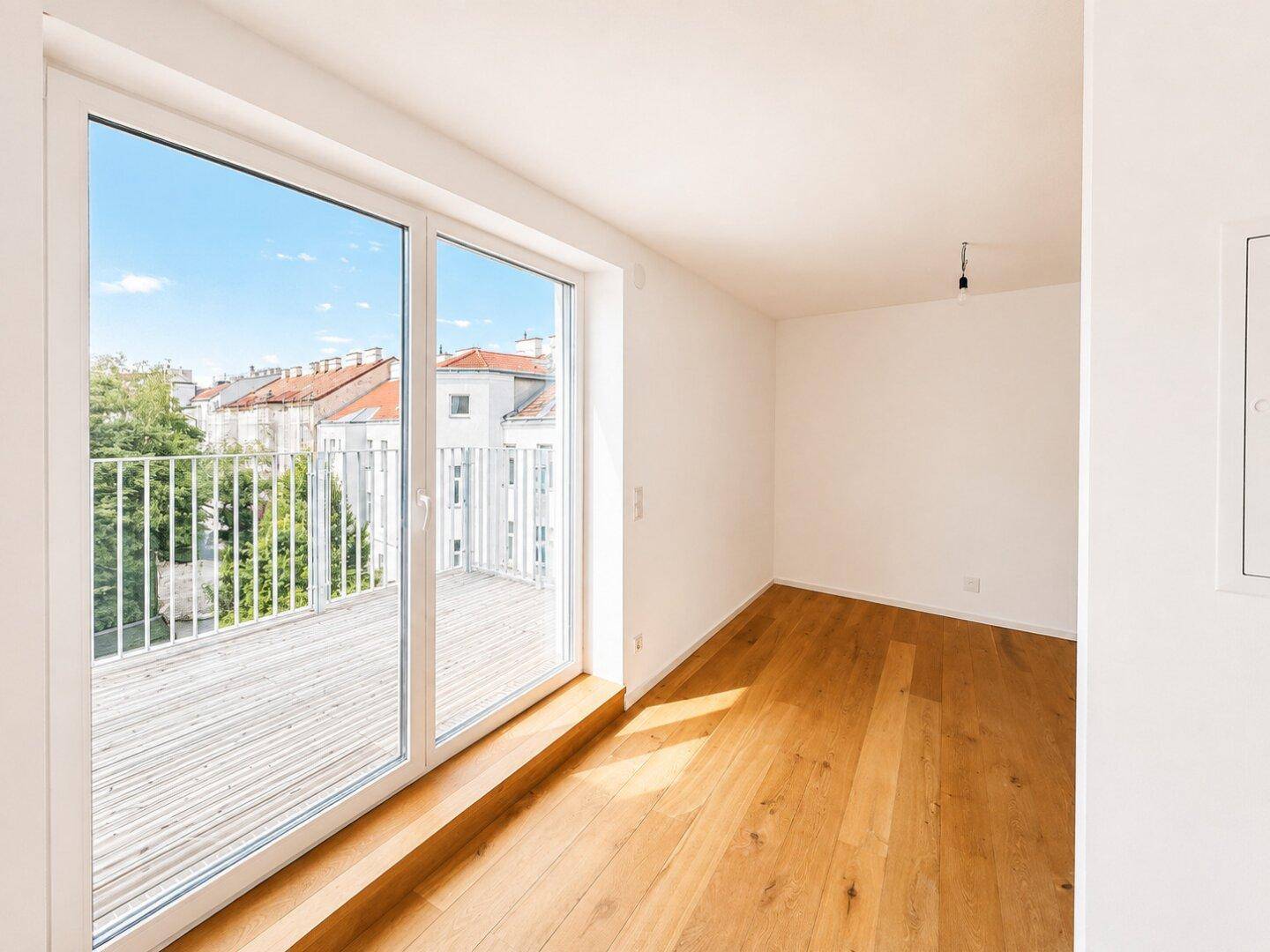 Heller Raum mit Holzboden und großen Schiebetüren, die auf einen sonnigen Balkon führen.