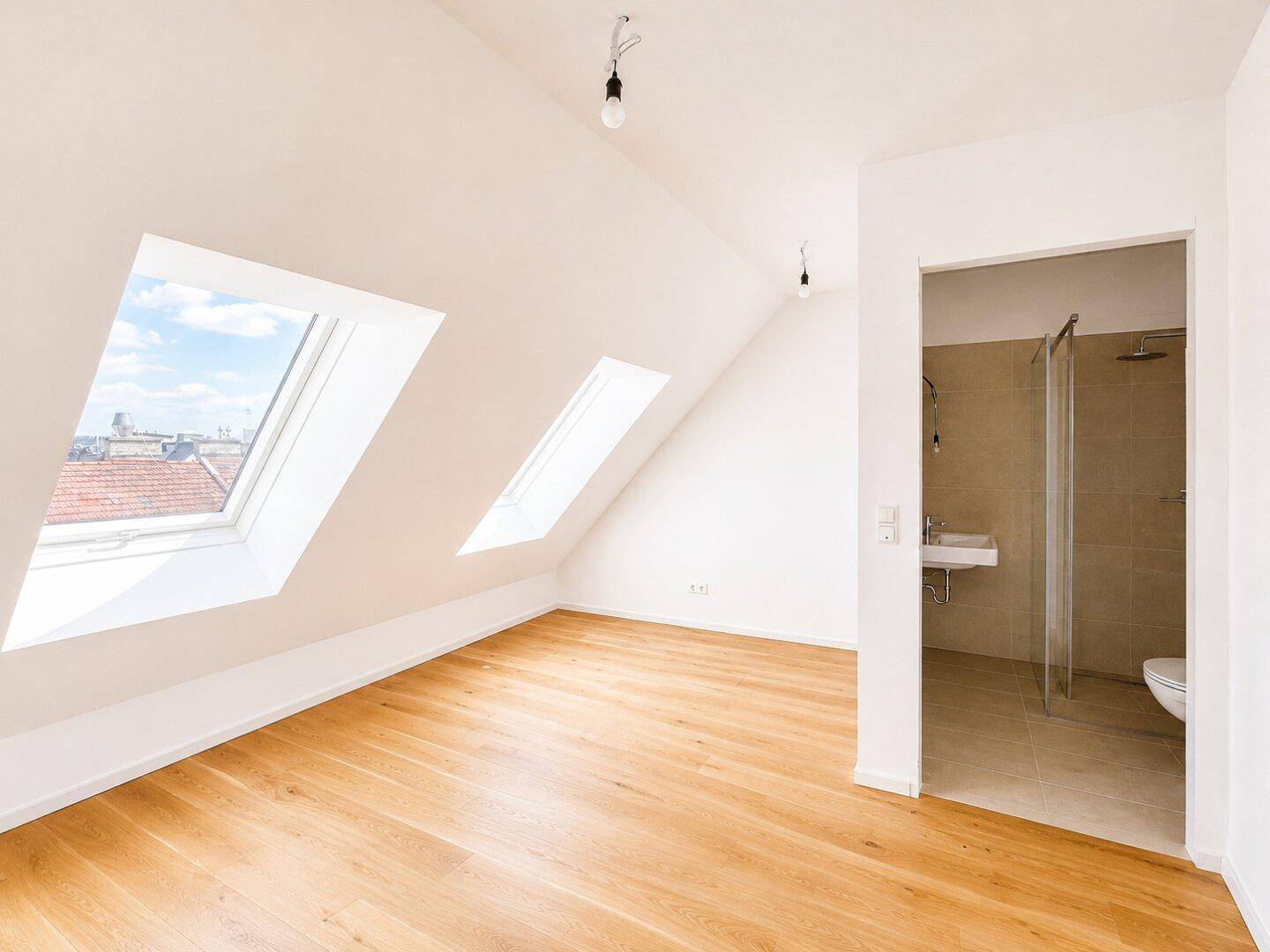 Heller Raum mit Dachschrägen, Holzboden, Dachfenstern und angrenzendem Badezimmer mit Dusche.