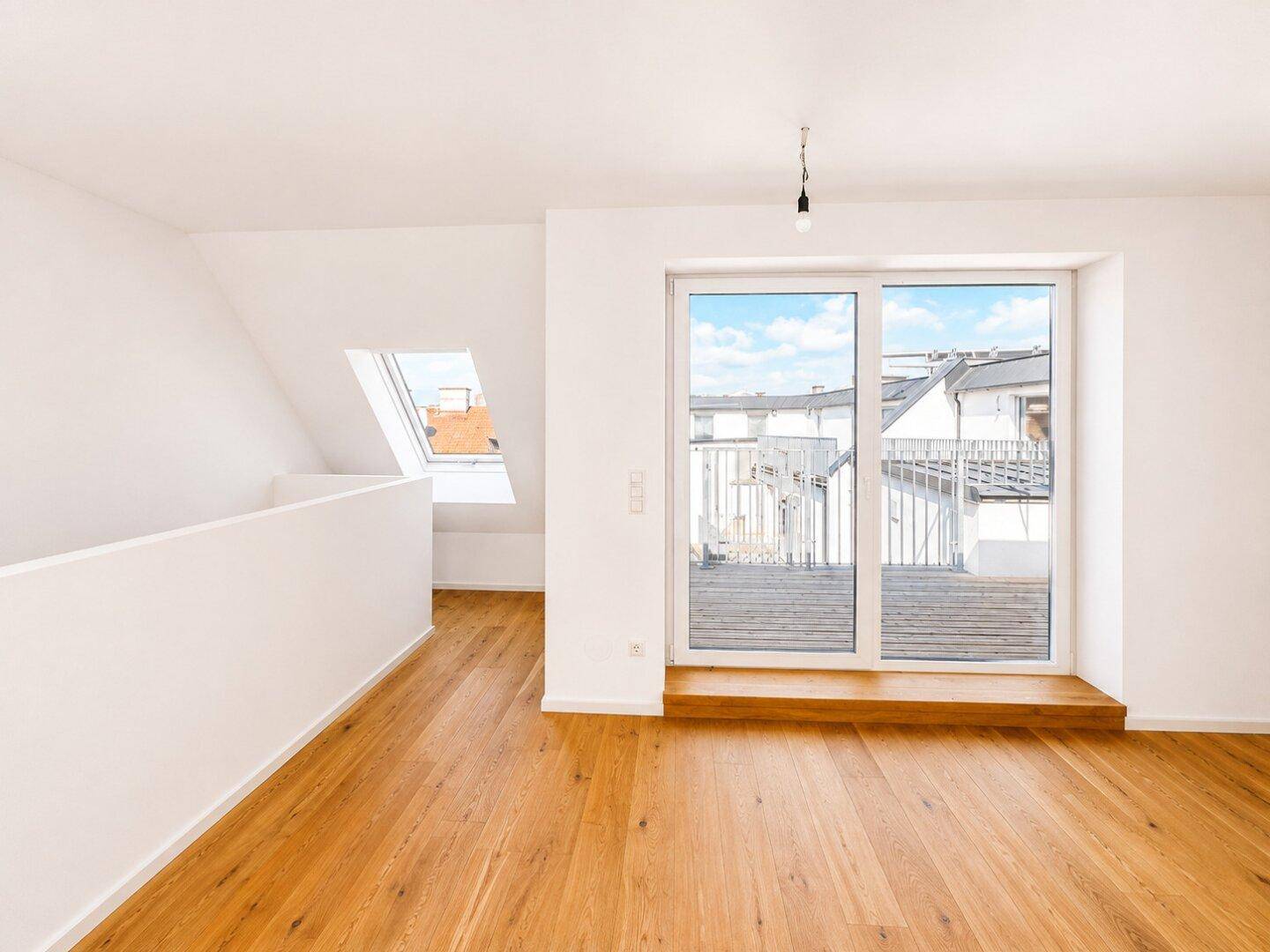 Geräumiger Raum mit Holzboden, Dachschrägen und Zugang zu einem Balkon sowie einem Dachfenster.