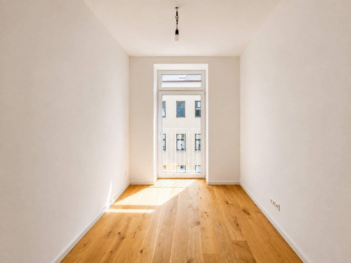 Schmaler Raum mit Parkettboden und einem französischen Balkon, der viel Licht spendet.