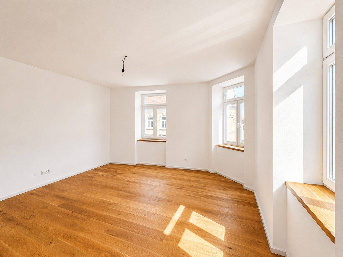 Geräumiges, helles Zimmer mit Parkettboden und mehreren Fenstern, ideal als Wohnbereich.
