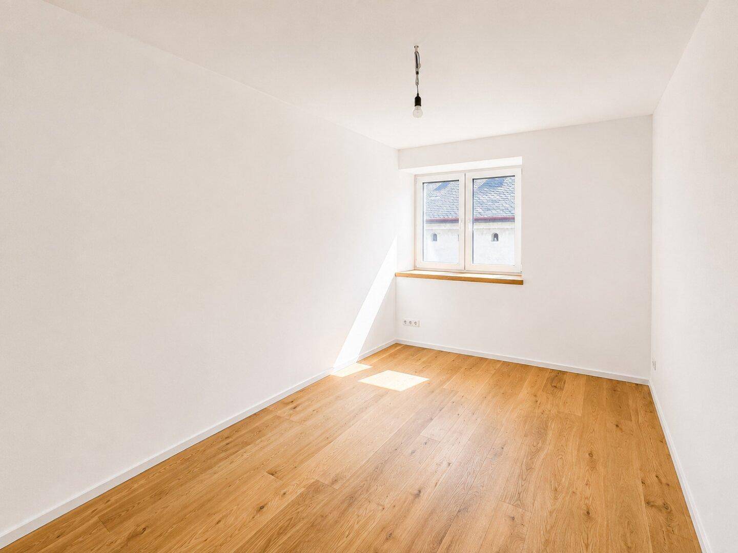 Sonniger, leerer Raum mit einem Fenster und Holzboden, ideal für ein Schlafzimmer oder Büro.