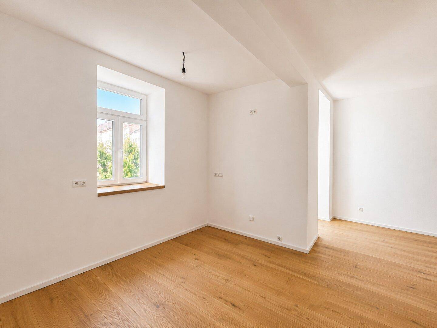 Leerer Raum mit Holzboden und einem großen Fenster, das viel Tageslicht hereinlässt.