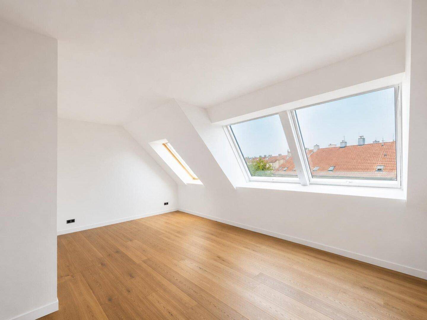 Leeres Zimmer mit Holzboden und Dachfenstern, die einen weiten Blick über die Dächer bieten.