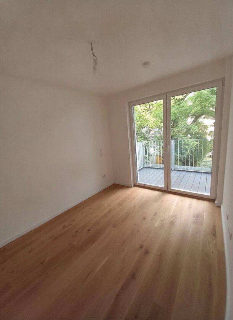 Leeres Zimmer mit Holzboden und großem Fenster, das Zugang zu einem Balkon bietet.