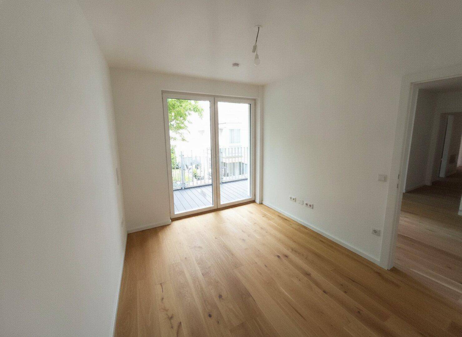 Helles, leeres Zimmer mit Holzboden und Zugang zu einem Balkon, ideal als Schlafzimmer.