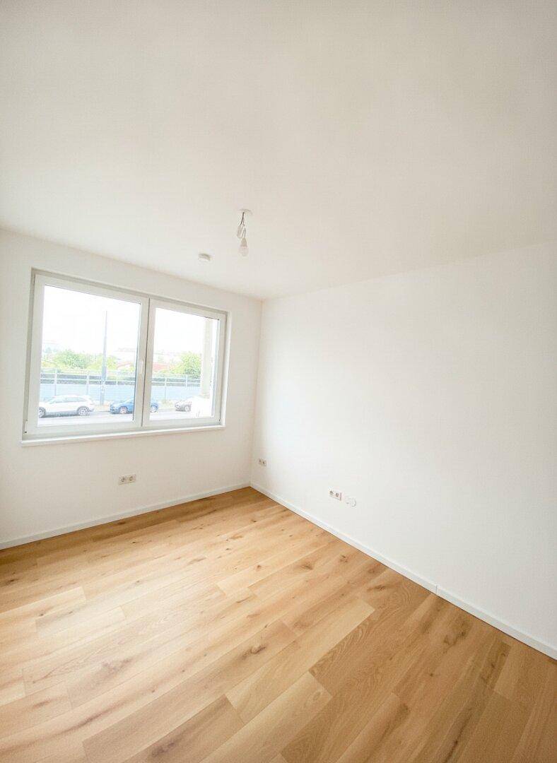 Leeres Zimmer mit Holzboden und großem Fenster, das viel Tageslicht hereinlässt.