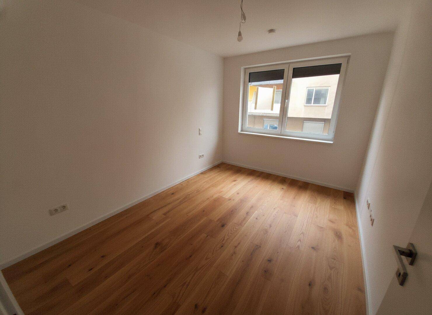 Helles Zimmer mit Holzboden und Fenster, das einen Blick auf die umliegenden Gebäude bietet.