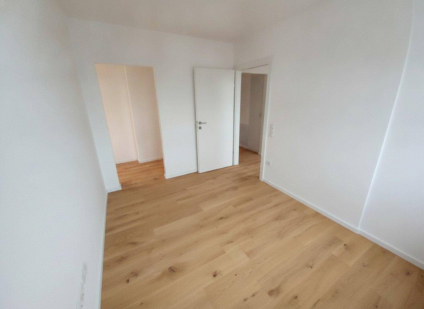 Leeres Zimmer mit Holzboden und weiß gestrichenen Wänden, mit Zugang zu weiteren Räumen.