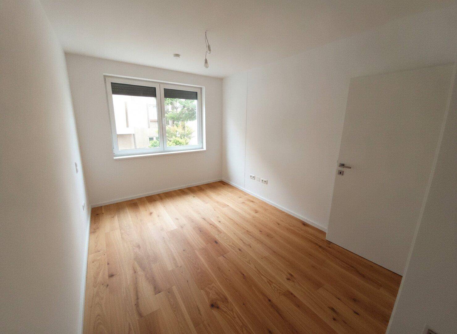 Leeres Zimmer mit Holzboden und Fenster, das viel Tageslicht hereinlässt und einen grünen Ausblick bietet.