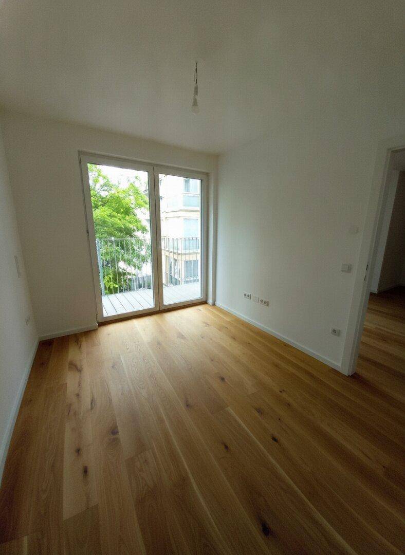 Leerer Raum mit hellem Holzparkett und großem Fenster mit Zugang zu einem Balkon.