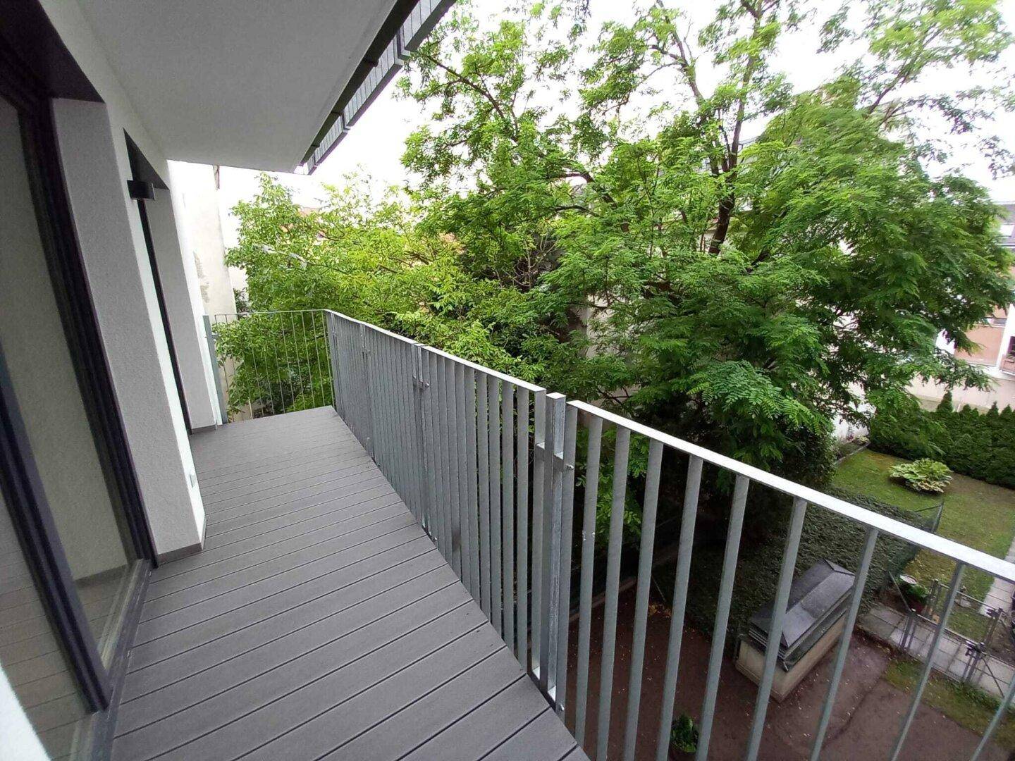 Balkon mit grauen Dielen und Metallgeländer, umgeben von Bäumen und einem Innenhof.