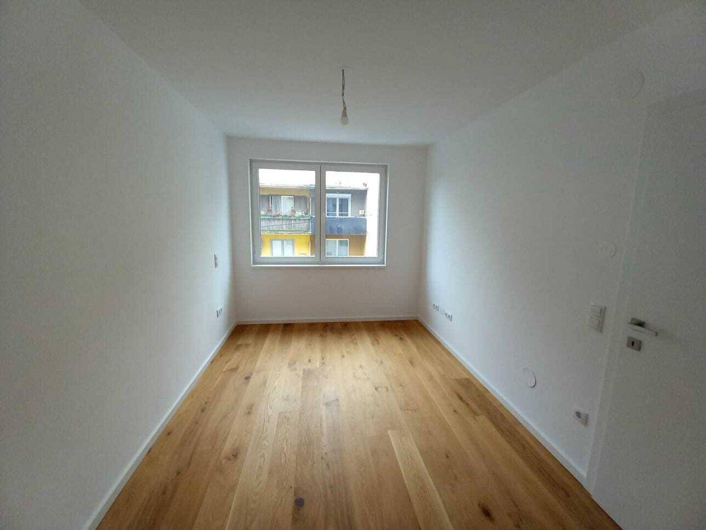Leeres Zimmer mit Holzboden und Fenster, das einen Blick auf umliegende Gebäude bietet.