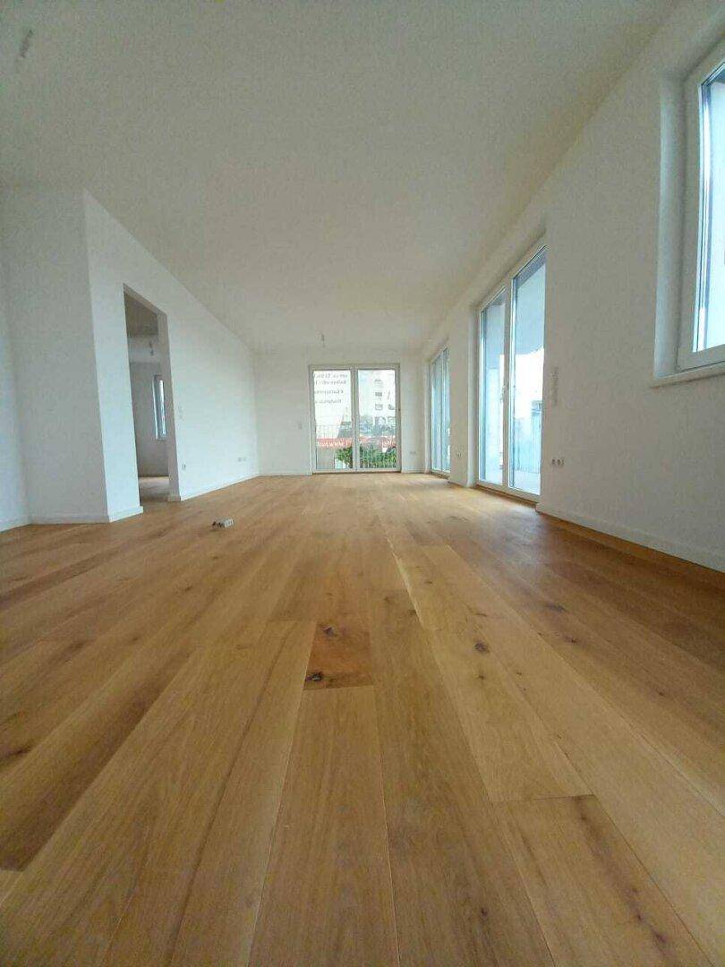 Heller, leerer Raum mit Holzdielenboden und großen Fenstern zum Balkon.