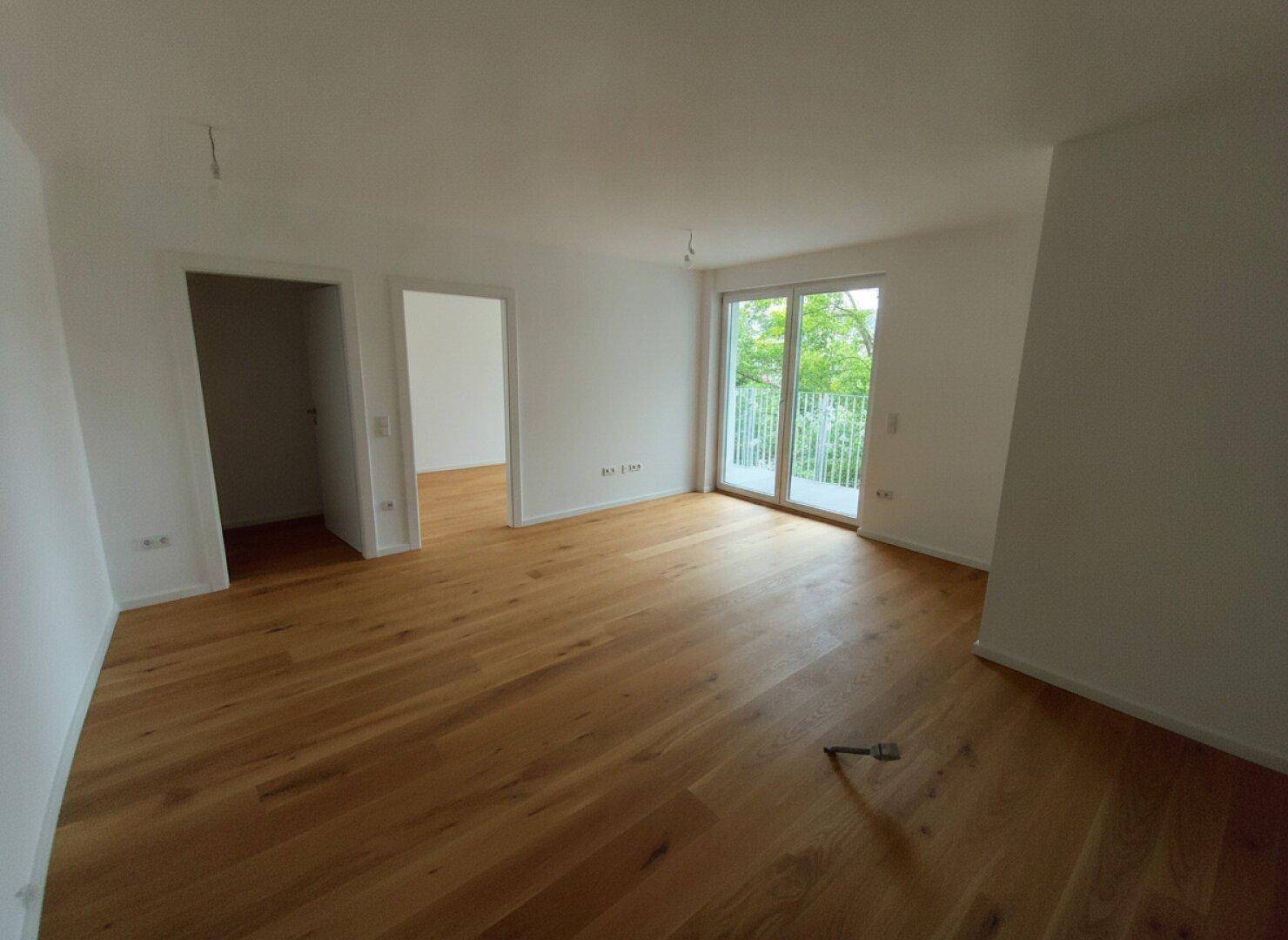 Unmöblierter Wohnbereich mit hellem Holzboden und Zugang zu einem Balkon mit grüner Aussicht.