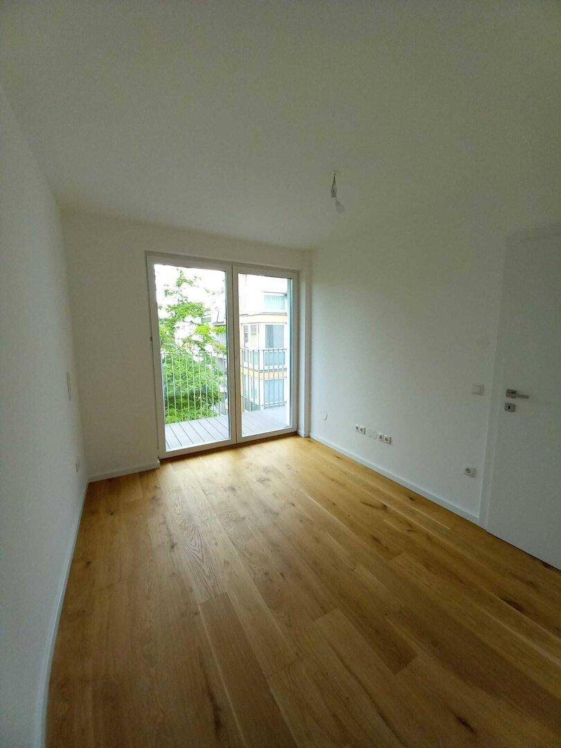 Unmöbliertes Zimmer mit Holzboden und Balkonzugang, bietet viel Tageslicht und frische Luft.