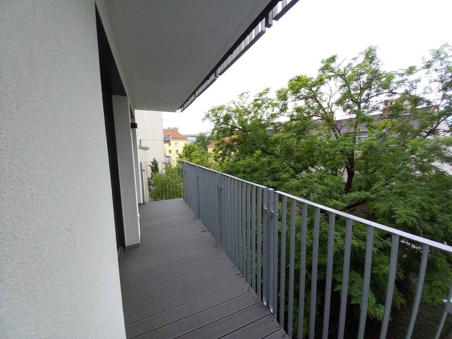 Langer Balkon mit grauem Bodenbelag und Metallgeländer, umgeben von grünen Bäumen.