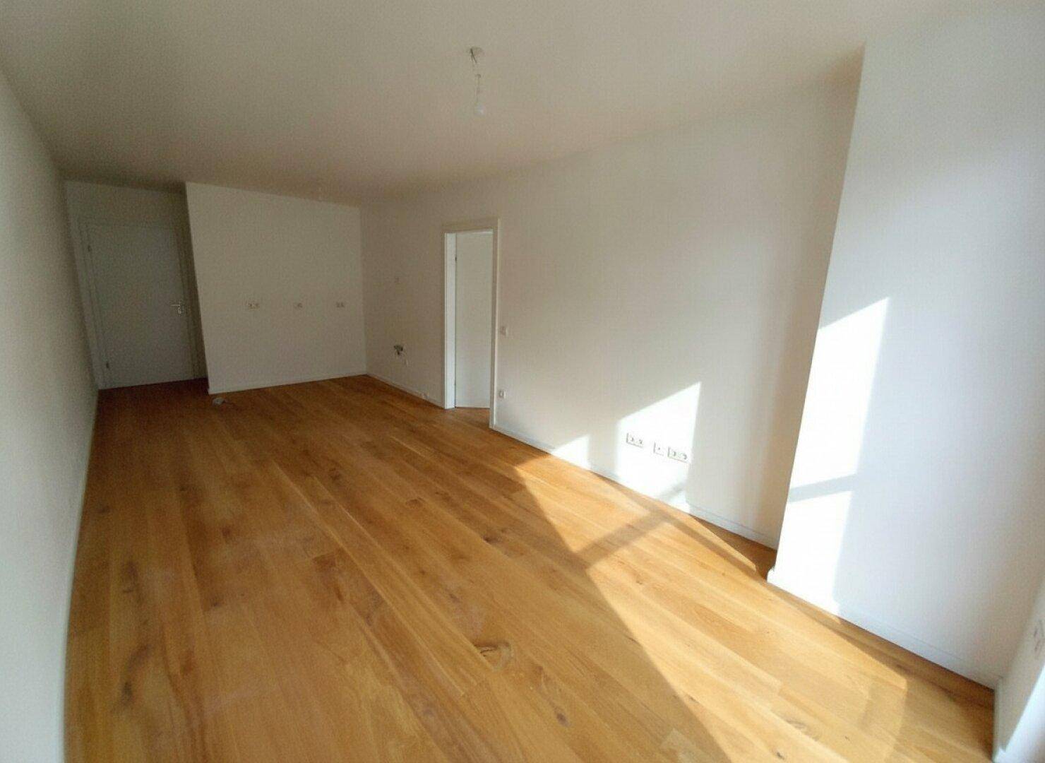 Leerer Raum mit hellem Holzfußboden und weißen Wänden, durchflutet von natürlichem Licht.