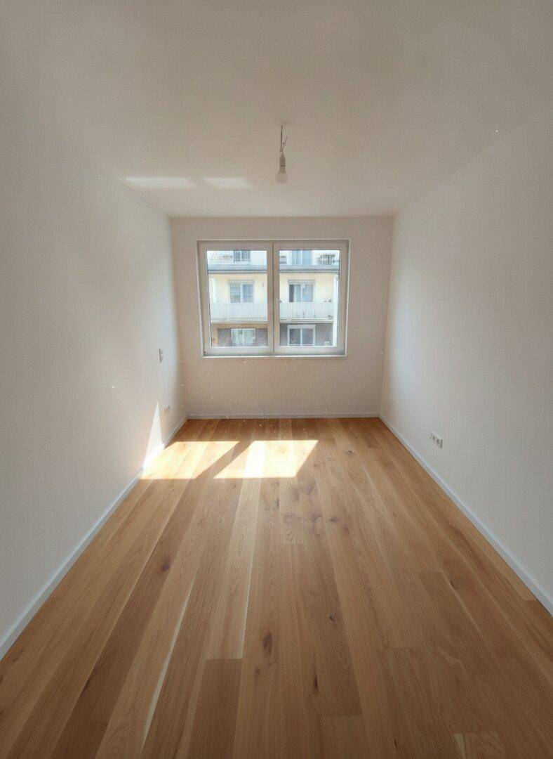 Leerer Raum mit hellem Holzfußboden und Fensterblick auf die umliegenden Gebäude, sonnendurchflutet.