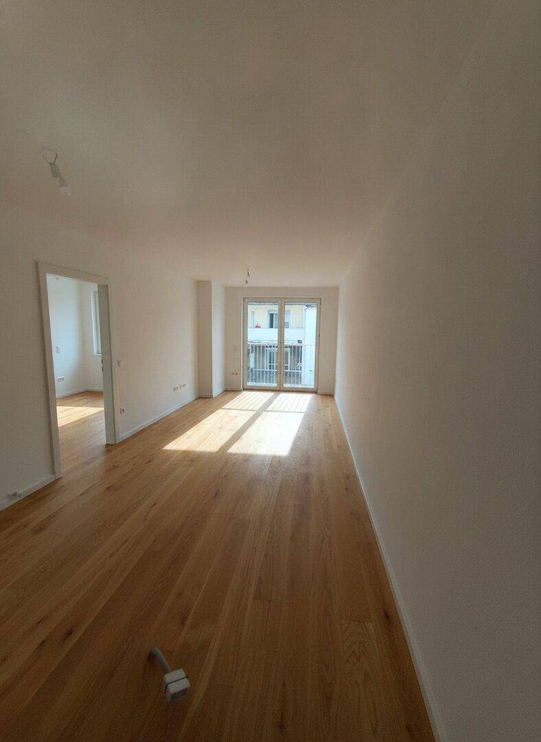 Heller, leerer Raum mit Holzfußboden und Zugang zu einem Balkon, gut beleuchtet durch Tageslicht.