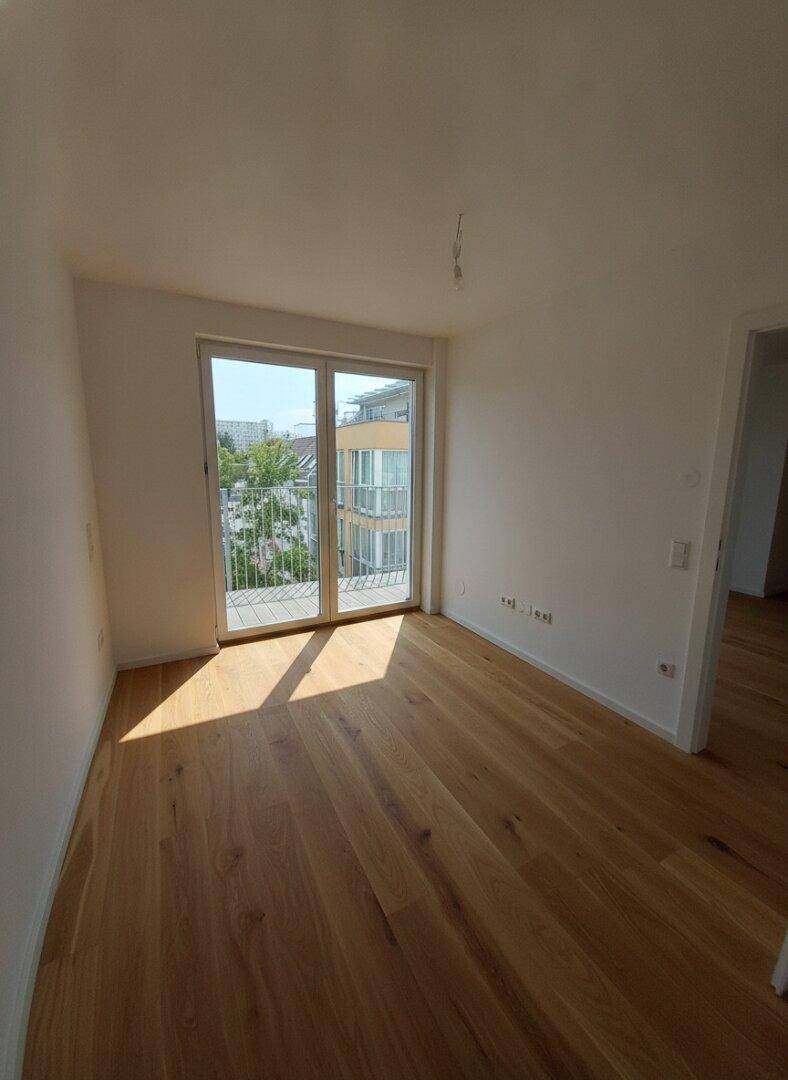 Heller, unmöblierter Raum mit Holzboden und großem Fenster, das viel Tageslicht hereinlässt.