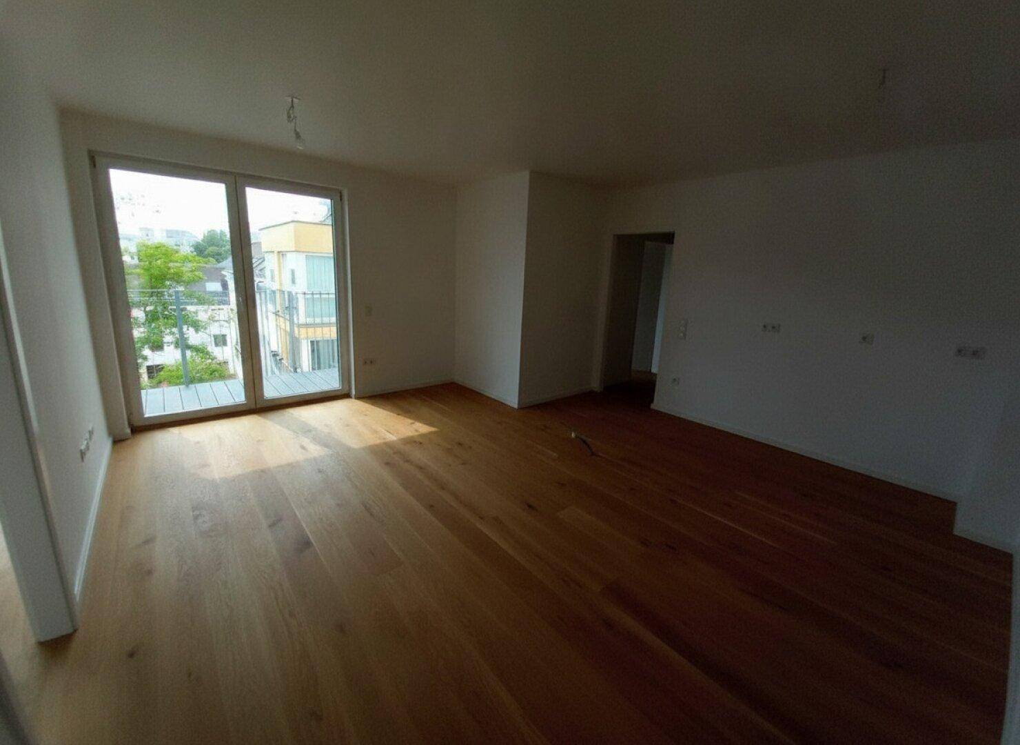 Geräumiger, unmöblierter Raum mit Holzboden und großen Fenstern, die viel Licht spenden.