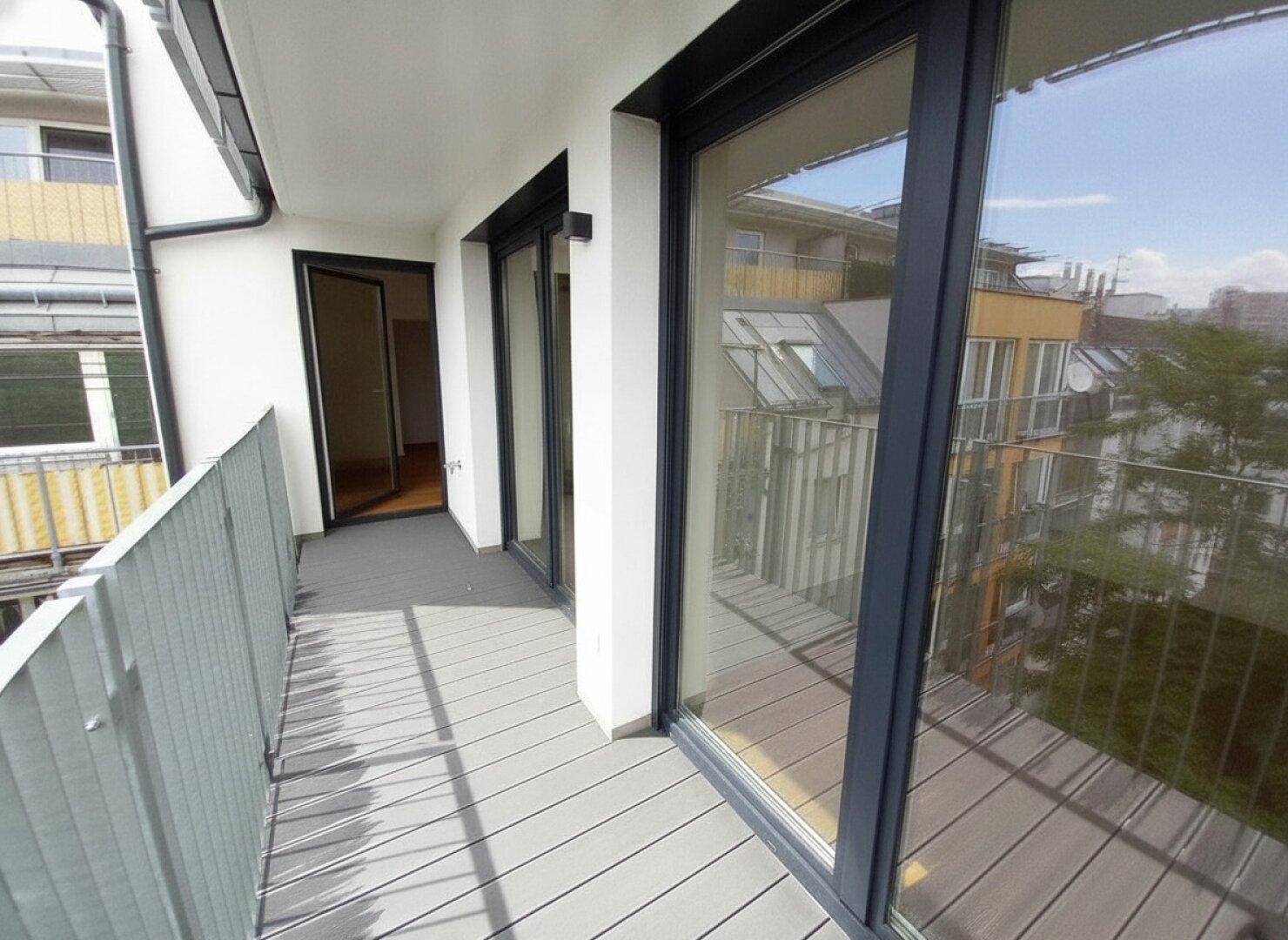 Langer Balkon mit grauem Bodenbelag und Blick auf die umliegenden Gebäude und Bäume.