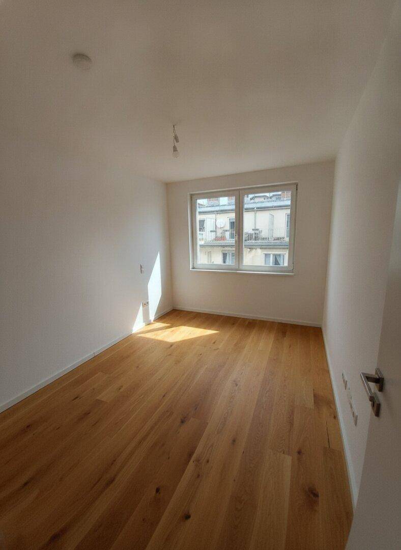 Leeres Zimmer mit Holzboden und Fenster, das auf die Nachbargebäude blickt und viel Licht hereinlässt.