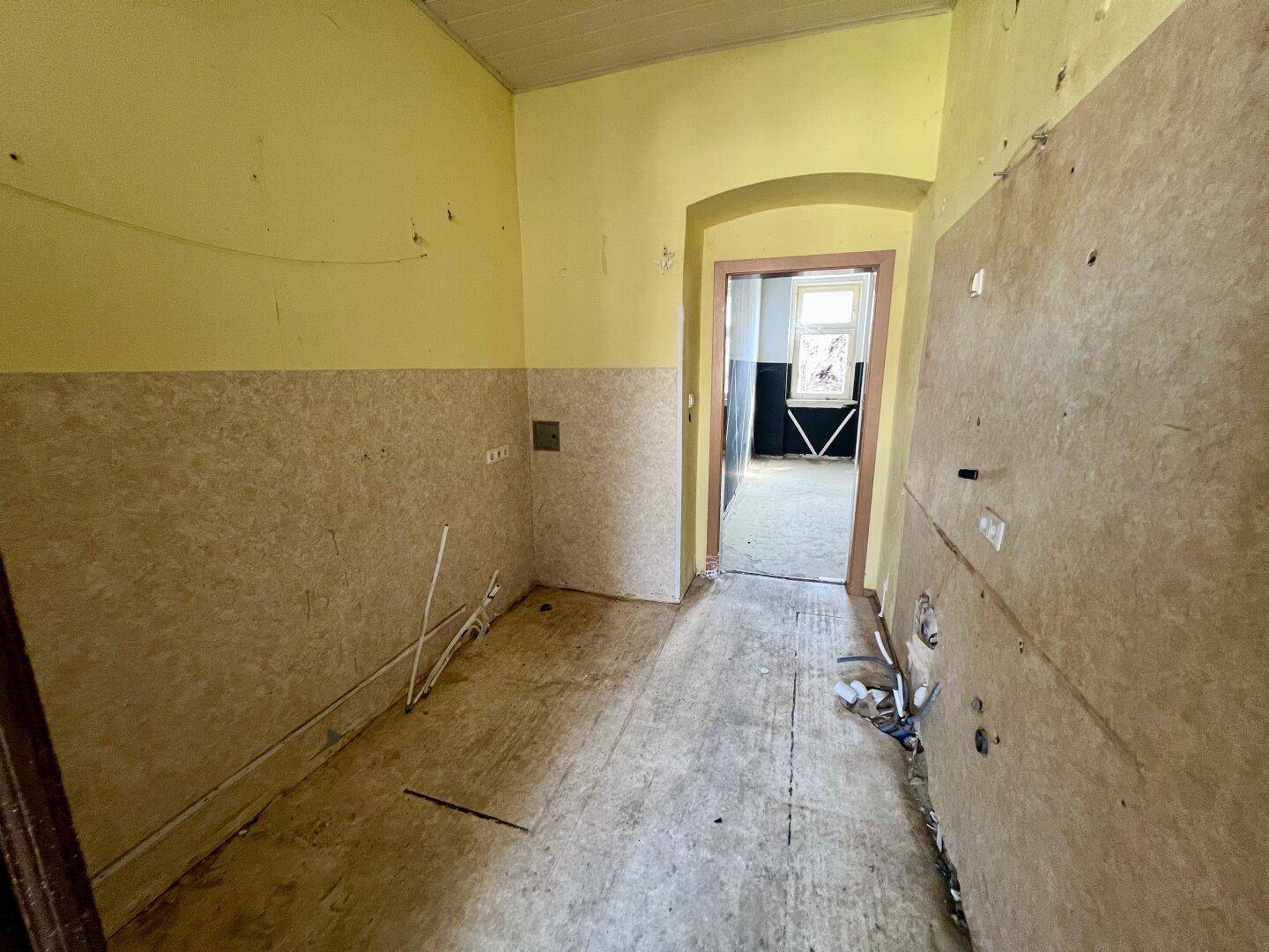 Flur oder Abstellraum in Renovierung mit gelben Wänden, sichtbaren Installationen und Blick in ein weiteres Zimmer.