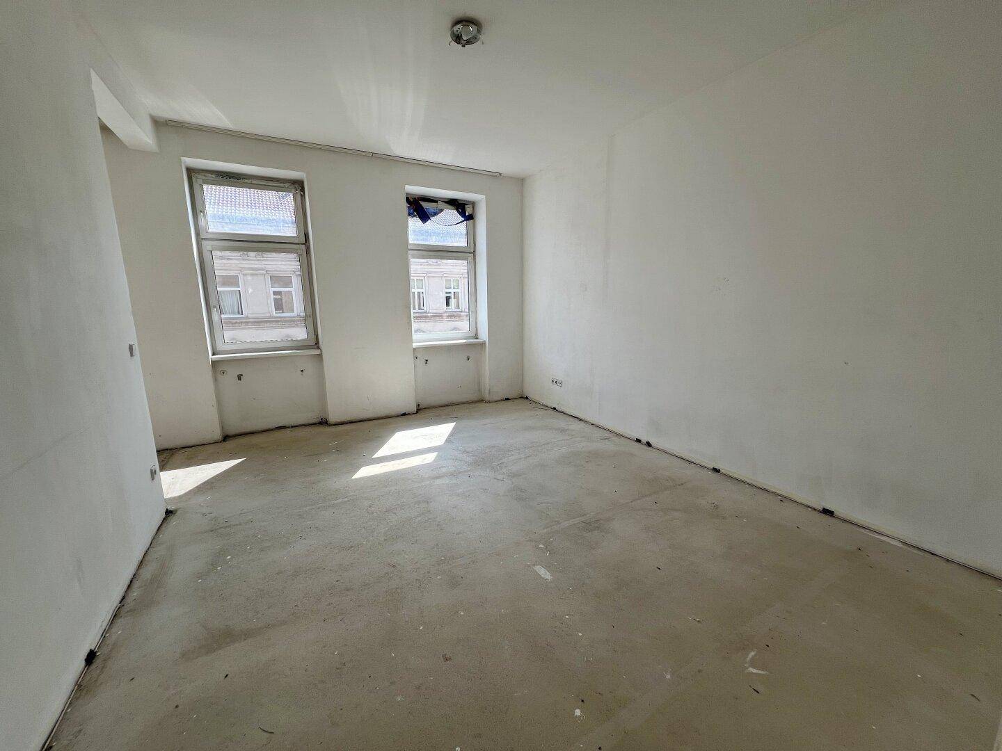 Heller, leerer Raum mit weißen Wänden und Betonboden, der viel natürliches Licht durch zwei Fenster erhält.