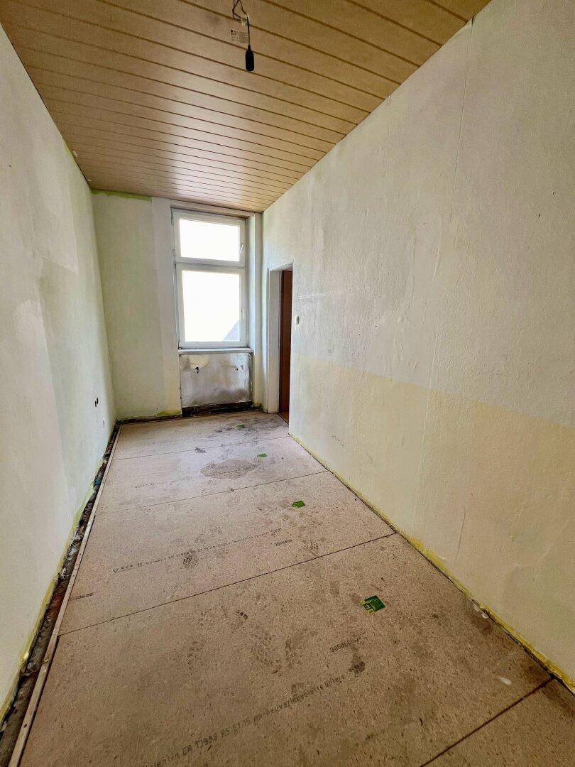 Schmaler, unmöblierter Raum mit Holzpaneeldecke und einem Fenster, teilweise grüne Wände.