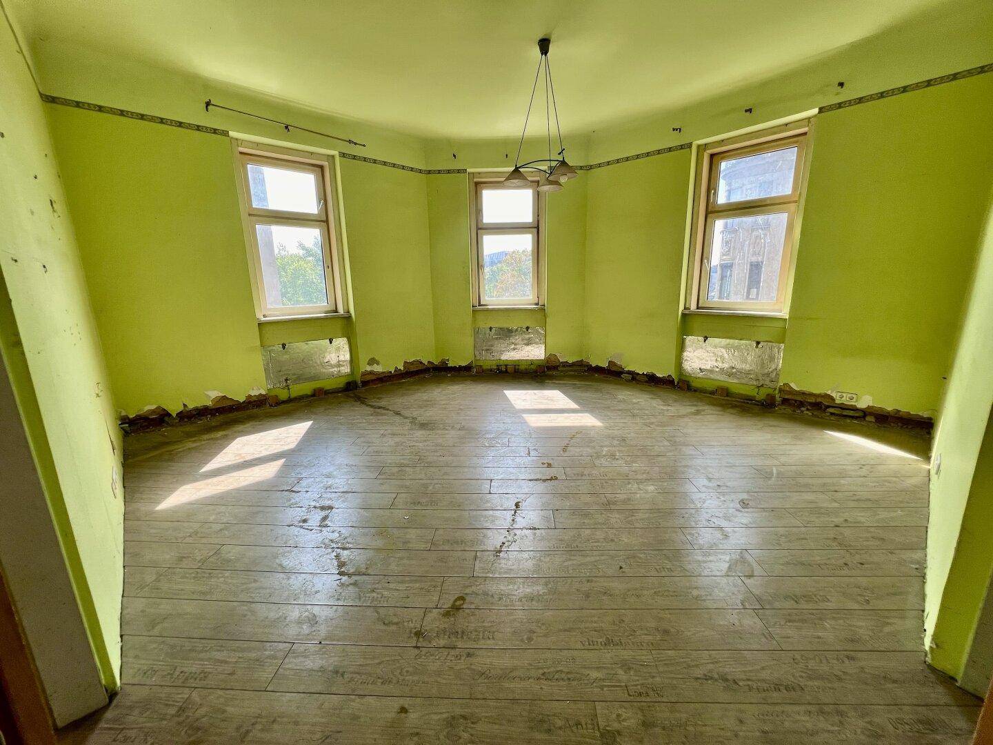 Eckzimmer mit drei Fenstern, leuchtend grünen Wänden und Laminatboden, renovierungsbedürftig.