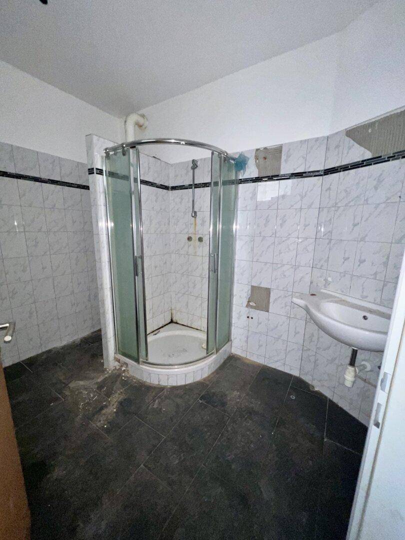 Badezimmer mit Duschkabine, Waschbecken und Fliesenboden, renovierungsbedürftig.