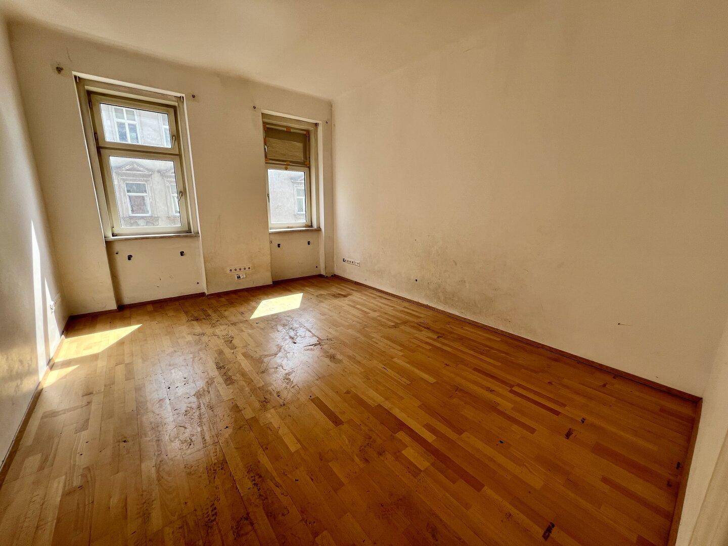 Heller Raum mit Holzboden und zwei Fenstern, der auf eine Renovierung wartet.
