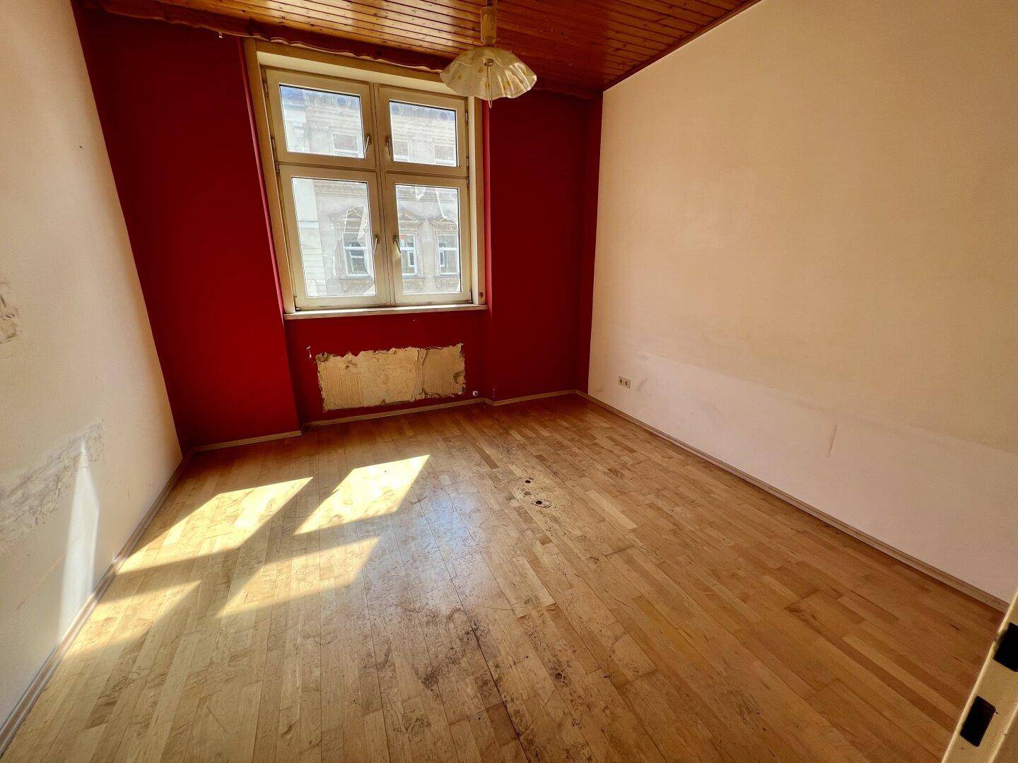 Heller Raum mit Holzbalkendecke, Holzboden und einem Fenster, das Tageslicht hereinlässt.