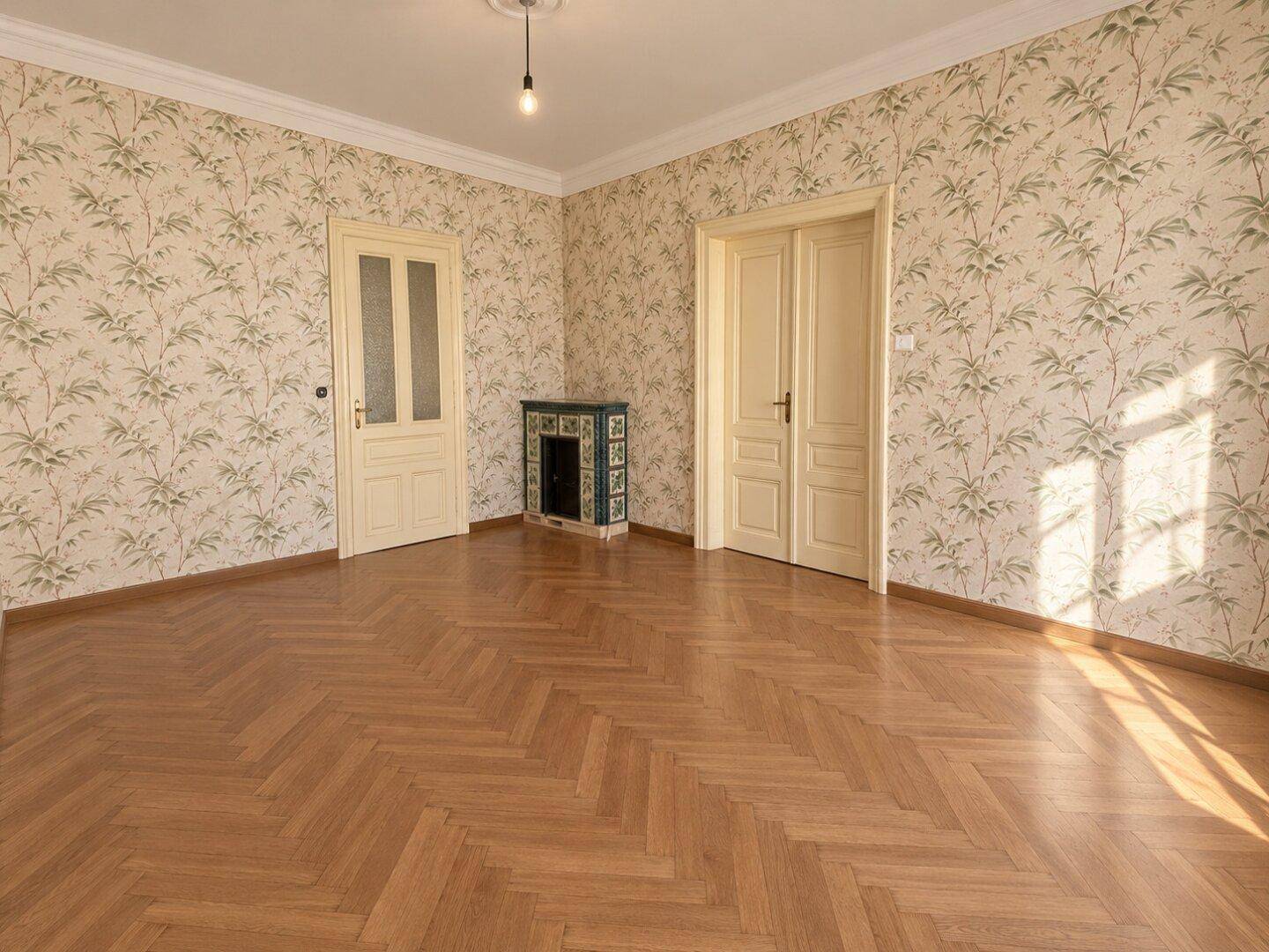 Geräumiges Zimmer mit Fischgrätparkett, floraler Tapete und einem traditionellen Kachelofen.