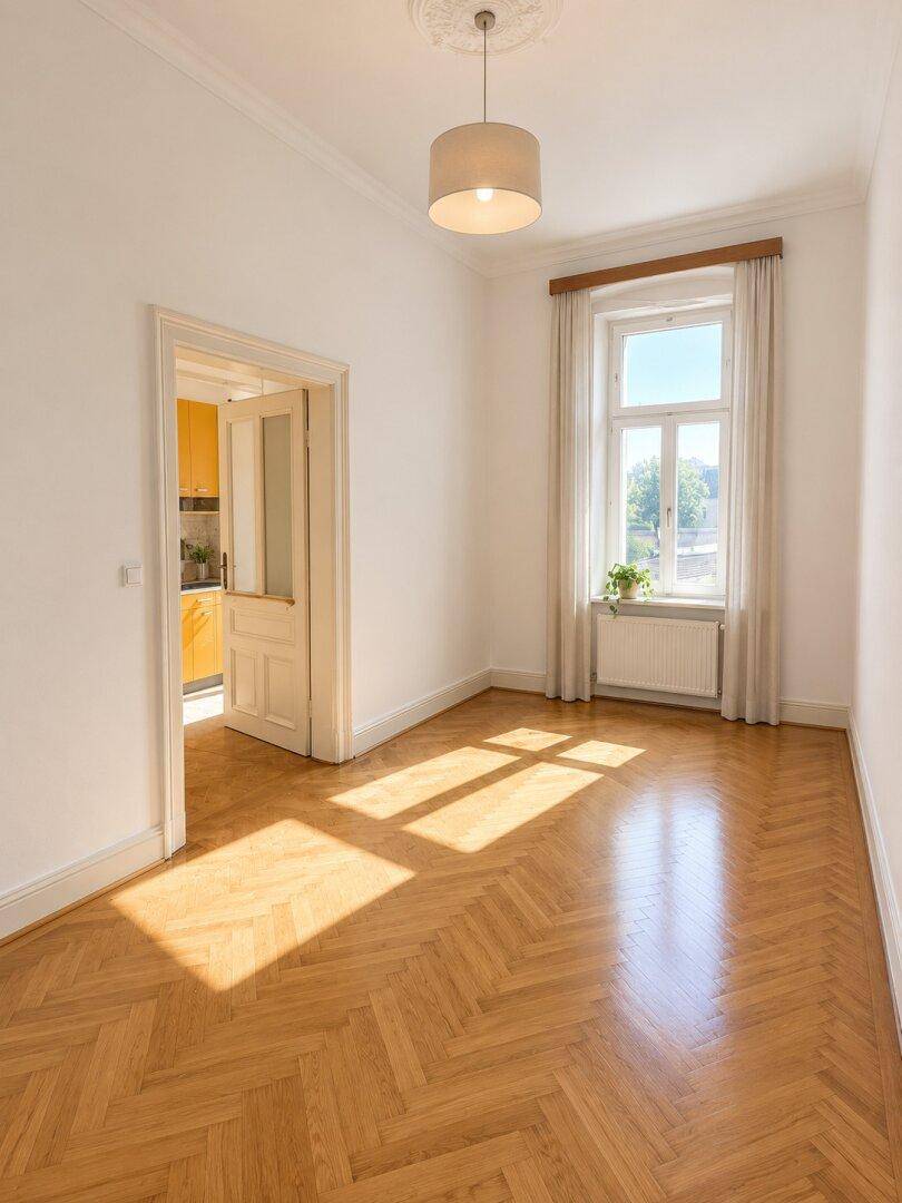 Helles Zimmer mit Fischgrätparkett, hohen Decken und Blick ins Grüne durch das Fenster.