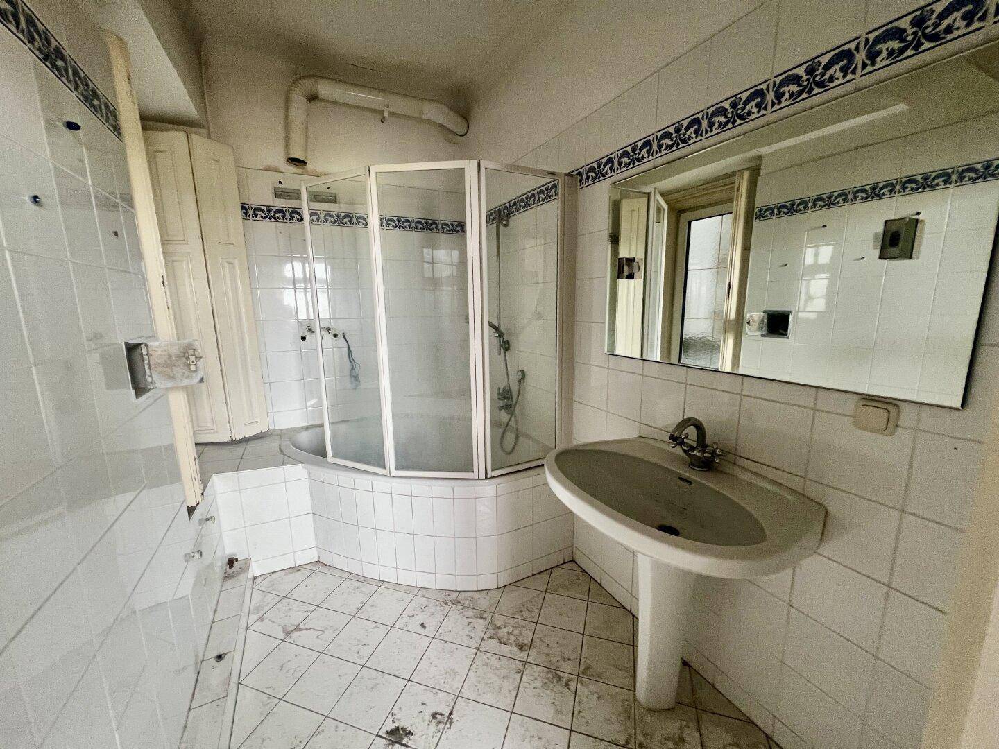 Veraltetes Badezimmer mit weißen Fliesen, blauer Bordüre, Eckdusche und Waschbecken.