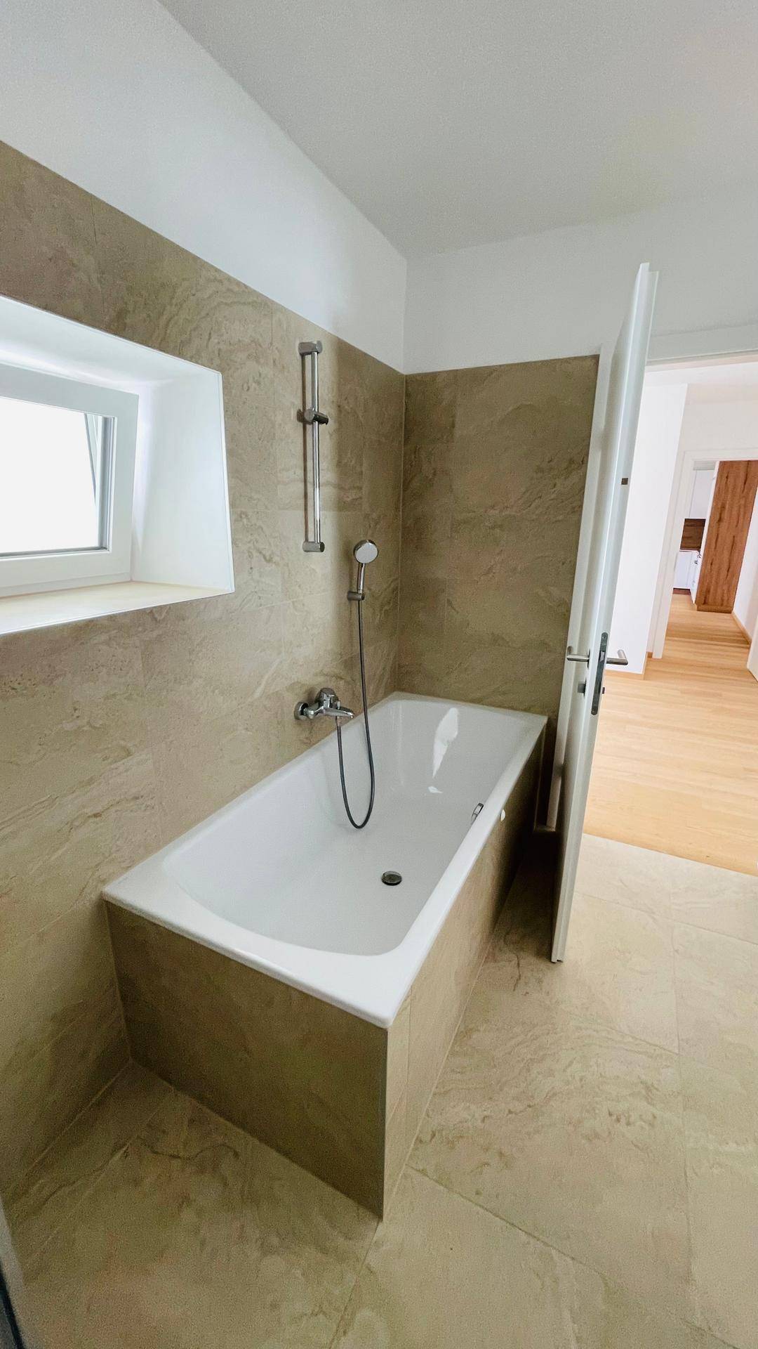 Badezimmer mit Badewanne, beigefarbenen Fliesen und einem Fenster, das Tageslicht hereinlässt.