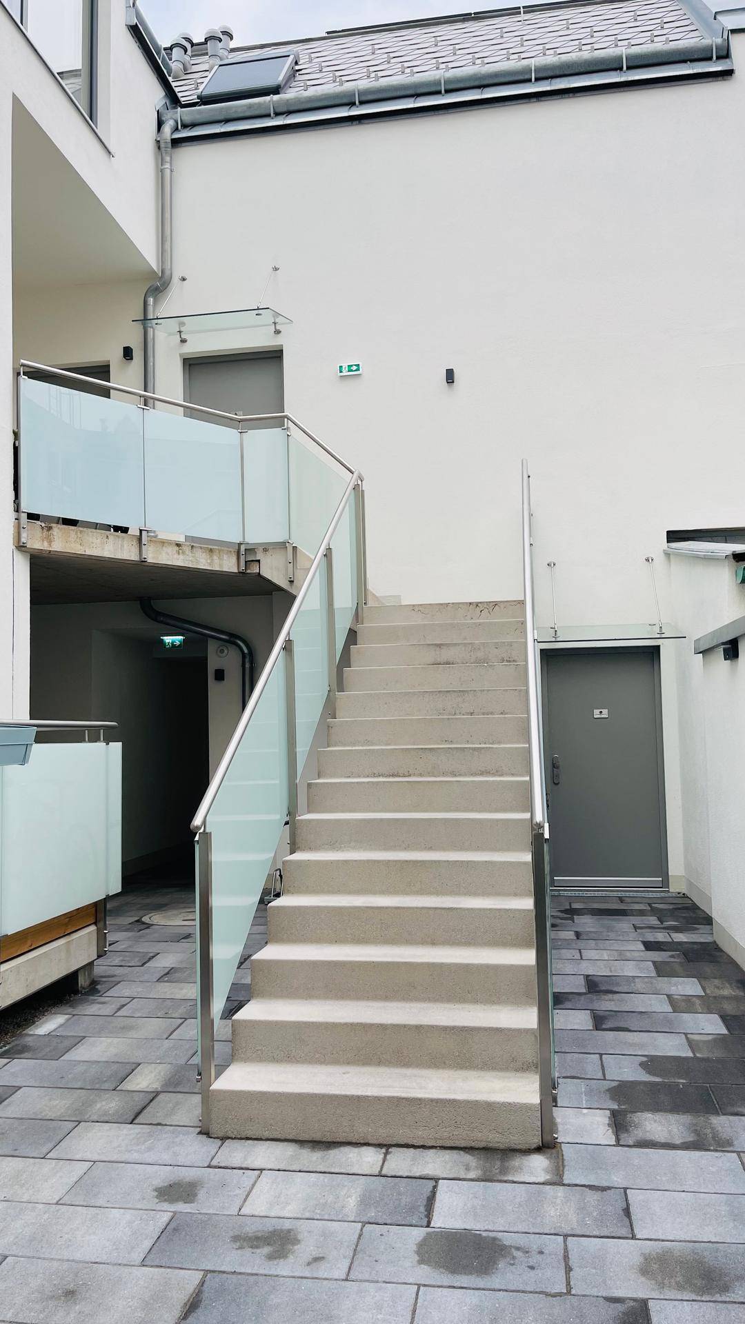 Außentreppe mit modernen Glasgeländern und gepflastertem Bereich vor dem Gebäudeeingang.
