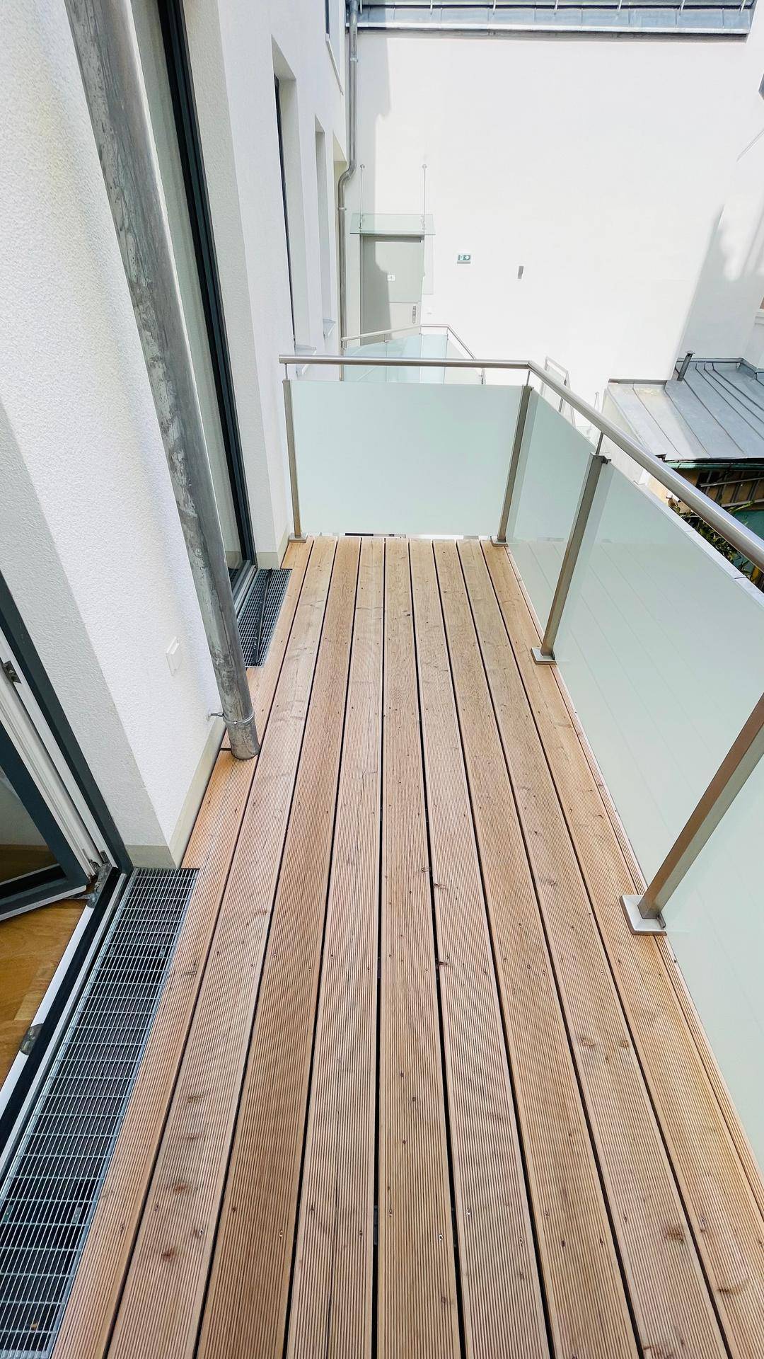 Schmaler Balkon mit Holzdielenboden und modernen Glasgeländern, ideal für Entspannung im Freien.