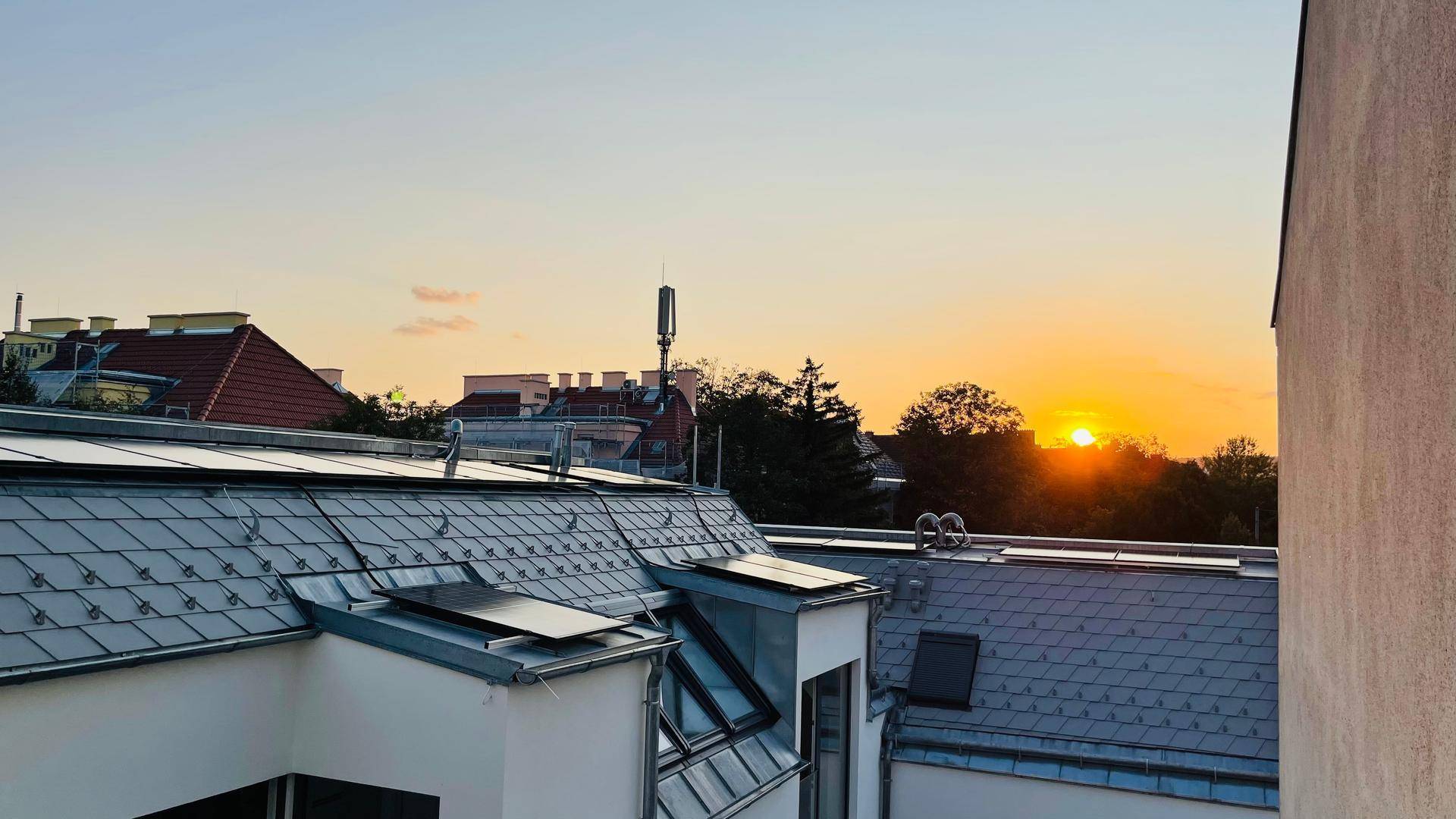Sonnenuntergang über den Dächern der Stadt mit sichtbaren Solarmodulen auf einem grauen Ziegeldach.