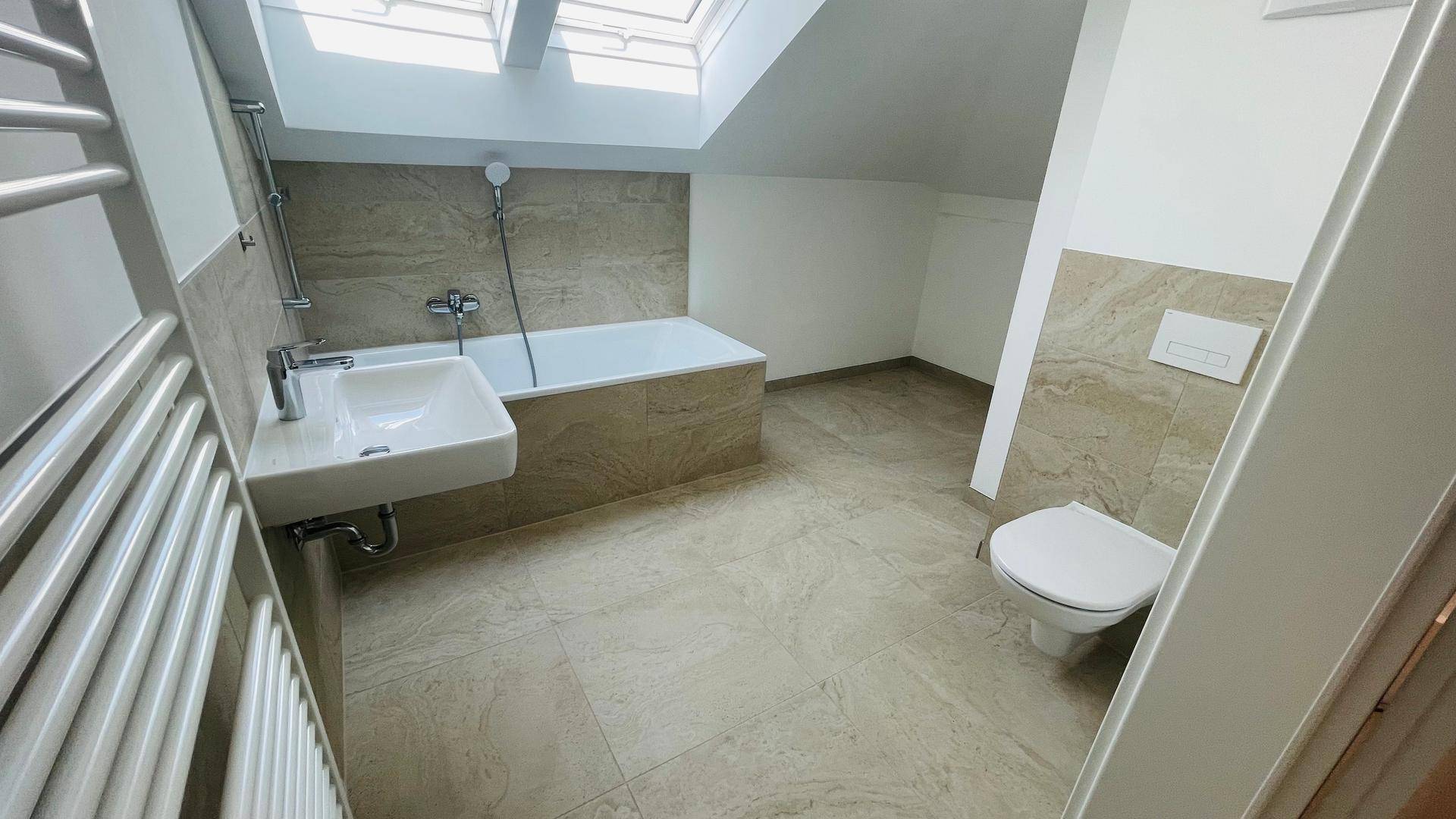 Modernes Badezimmer mit Badewanne, Waschbecken und WC unter einem Dachfenster in der Schräge.